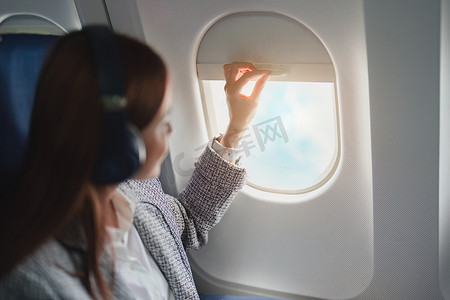 一位成功的亚洲女商人或企业家在飞机上穿着正式的西装，坐在商务舱里，在飞行过程中透过窗户看，听音乐