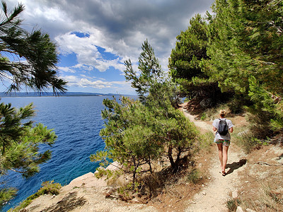 年轻活跃的 feamle 游客背着小背包走在松树间的沿海小路上，寻找偏远的海湾，在克罗地亚的海边安静地游泳。