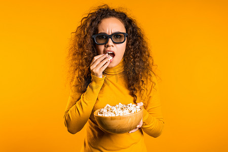 戴 3D 眼镜的年轻女性在黄色背景下看电影和吃爆米花