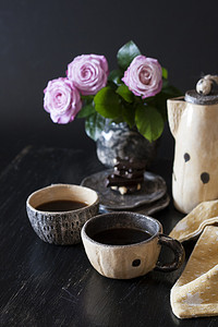 黑茶壶摄影照片_两杯黑咖啡、黑巧克力、一个黄色茶壶和一个深色背景上插着粉红玫瑰的花瓶。