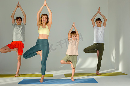一个漂亮苗条的女人、一个男孩和两个青少年做瑜伽练习，站在垫子上摆出树姿