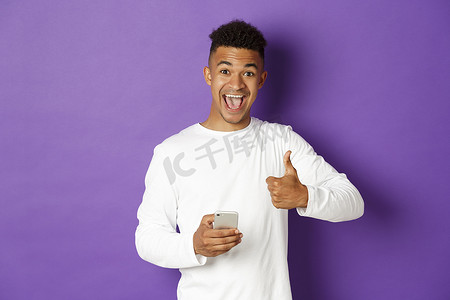 橘黄紫色摄影照片_一张惊讶而满意的非洲裔美国人的照片，他拿着手机，竖起大拇指表示赞同，就像什么东西一样，站在紫色背景上