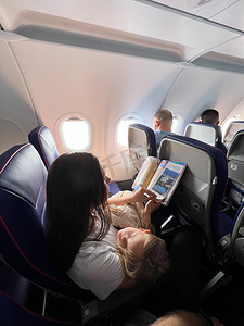 在飞机上睡觉摄影照片_抱着熟睡女儿的妈妈坐在飞机座位上看杂志