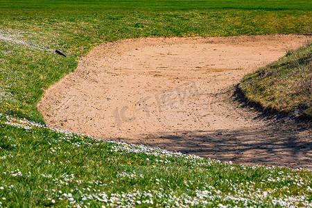 沙坑摄影照片_德国一个高尔夫球场上有一个带耙子的沙坑，春天沙子和草坪上种满了雏菊