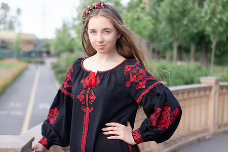 刺绣服装摄影照片_迷人的乌克兰年轻女子在户外穿着民族红色和黑色刺绣连衣裙。