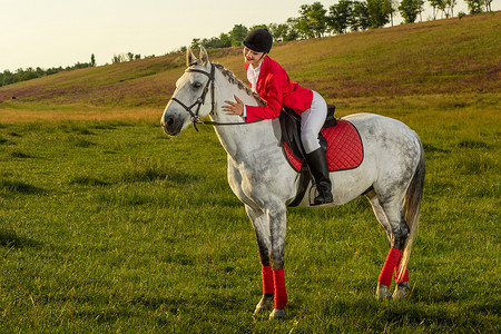 夕阳下的人摄影照片_年轻的女骑手，穿着红色的 redingote 和白色的马裤，她的马在夕阳的余晖下。