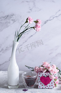 白色花瓶里的粉色康乃馨和心形粉色盒子里的咖啡杯，