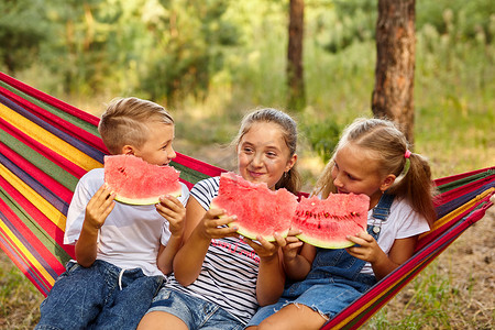 开玩笑的摄影照片_孩子们坐在色彩缤纷的吊床上，在户外吃西瓜，开玩笑。