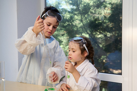 化学实验室穿着摄影照片_可爱的孩子们，穿着白色实验室外套和安全护目镜的男孩和女孩，在化学课上做科学实验
