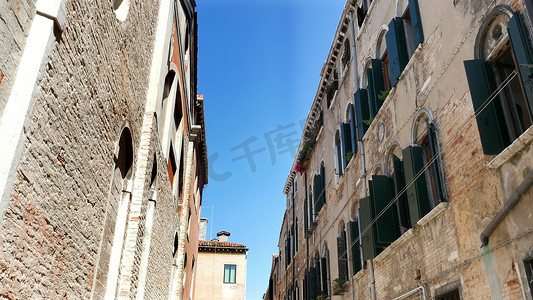 窗蓝天摄影照片_意大利威尼斯 — 2018年7月7日：威尼斯美丽的老建筑，一栋房子，窗台上有旧的绿色百叶窗和鲜花，在蓝天的衬托下，在炎热的夏日，