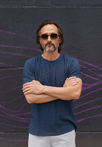 英俊成熟男子的肖像站在黑墙旁边，双手交叉，戴着墨镜、深蓝色 T 恤和浅色短裤。