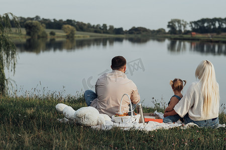年轻家庭野餐的后视图，爸爸妈妈带着他们的小女儿坐在城外的湖边