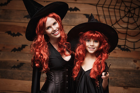美丽的白人母亲和她的女儿，长着红色的长发，穿着女巫服装和魔杖庆祝万圣节，在木制工作室背景上摆出蝙蝠和蜘蛛网的姿势