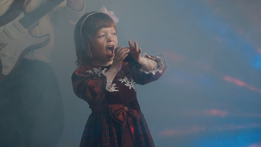 家庭舞台摄影照片_穿着复古裙子的小女孩在舞台上唱歌，她的父亲弹着电吉他