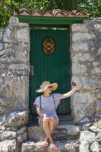 质感大摄影照片_美丽的年轻女游客戴着大草帽，自拍自拍，坐在地中海老城的白色复古木门和质感石墙前
