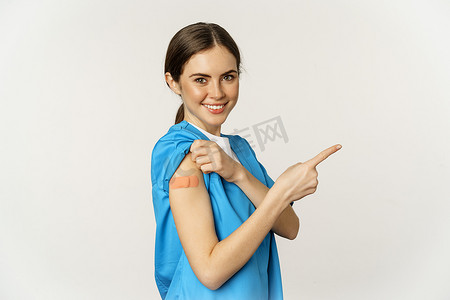 微笑的护士，穿着磨砂膏的女医护人员，手指向右，在肩上展示贴片，疫苗接种运动，covid-19 和流行病概念，白色背景