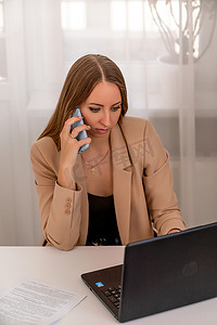 桌位摄影照片_欧洲职业女性坐在家庭办公室的桌子旁，拿着笔记本电脑，一位积极的女性一边在电脑上工作一边学习。