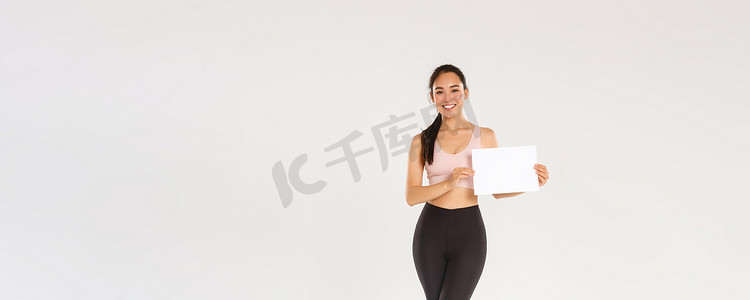全长微笑的漂亮亚洲黑发女运动员、穿着运动服的女运动员在白纸上展示标志、广告健身房会员资格或健身器材特价