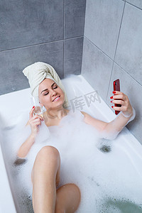 美女浴缸洗澡摄影照片_一位头戴毛巾的美女躺在白色的浴缸里，端着一杯香槟，正在打电话自拍。