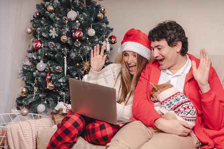 一对可爱的丈夫和妻子的肖像，戴着红色圣诞帽的笔记本电脑，与穿着毛衣的小狗进行视频通话，在家里享受圣诞节。