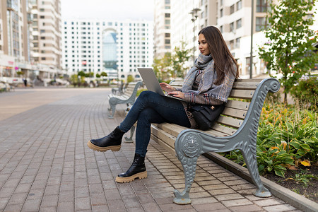 年轻女性坐在城市公园里，在住宅楼、概念自由职业和远程工作的背景下在线使用笔记本电脑工作