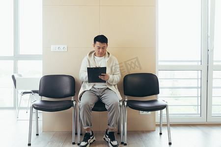 年轻的亚洲男性在候诊室接受采访或开会，坐在椅子上拿着纸，在办公室填写简历调查问卷