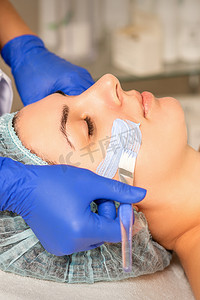 清洁面膜摄影照片_在水疗美容院的皮肤护理过程中，美容师用保湿清洁面膜覆盖女性面部皮肤。
