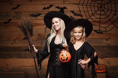 万圣节概念-美丽的白种人母亲和她的女儿穿着女巫服装庆祝万圣节，在木制工作室背景下分享万圣节糖果和糖果，在蝙蝠和蜘蛛网上。