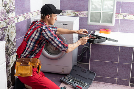 洗衣工摄影照片_穿着工作服的水管工正在修理房子里的洗衣机。