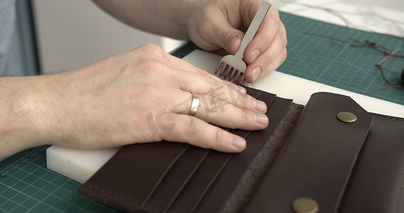 男人的手拿着锤子和打孔器，在他的工作室里打孔缝制皮革钱包。