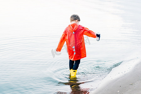 穿着红色雨衣和黄色橡胶靴的男孩在海滩玩水。