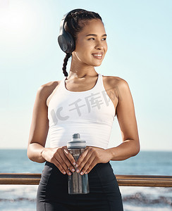 快乐的西班牙裔年轻女运动员一边戴着耳机听音乐，一边拿着水瓶微笑，一边在海滨长廊进行有氧运动，一边休息