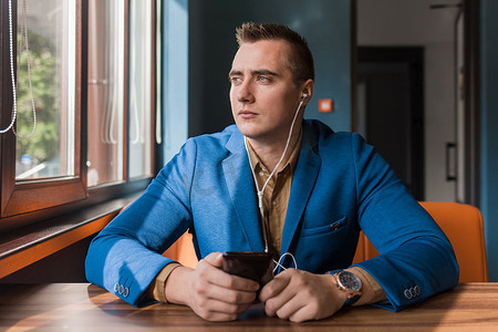 商人，一个穿着西装的白人外貌肖像的时尚帅哥，花时间在智能手机或手机上，坐在咖啡馆的桌子旁，望着窗外的背景