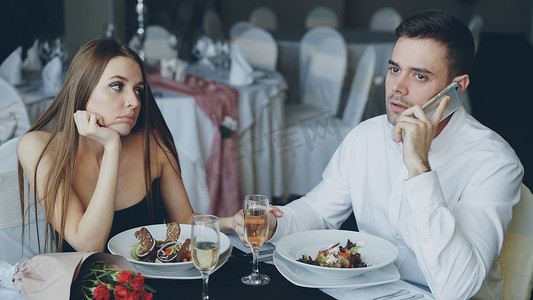 忙碌的年轻人正在打电话，而他的女朋友则坐在餐厅的餐桌旁感到无聊。