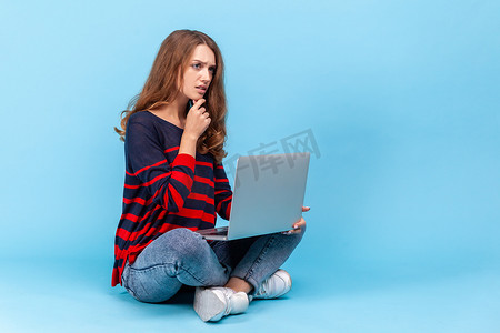 女人盘腿坐着，膝盖上拿着笔记本电脑，在线工作，表情若有所思。