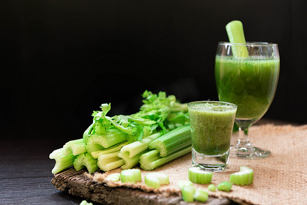 芹菜汁摄影照片_将芹菜汁混合在迎宾饮料杯中，并在木桌上放上一堆新鲜芹菜茎，叶子为黑色背景。