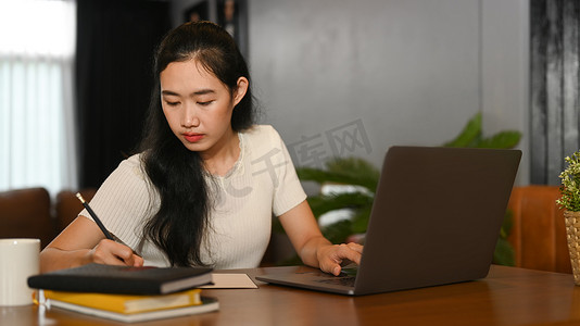 年轻的亚洲女士在笔记本电脑上在线学习，并在笔记本上写重要信息。
