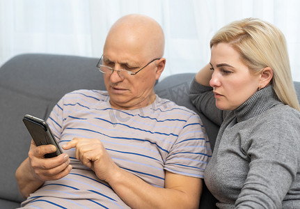 微笑的年长父亲与成年女儿一起使用手机，玩得开心，在社交网络中观看视频或在线购物，有爱心的年轻女性教成熟的父亲或祖父使用智能手机