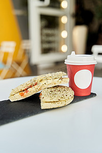 一家咖啡店的桌子上放着一个三明治，上面放着一杯红色塑料咖啡。