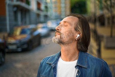 享受生活，闭着眼睛听音乐，站在户外老城街道上，英俊成熟的灰胡子男子，戴着无线耳机，穿着休闲装。