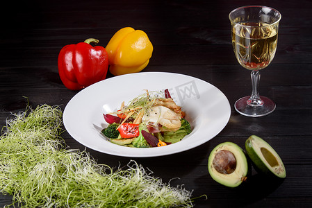 鸡柳配白酱和烤蔬菜，在白盘上装饰着微绿，背景是木桌上的一杯白葡萄酒、甜椒和鳄梨。