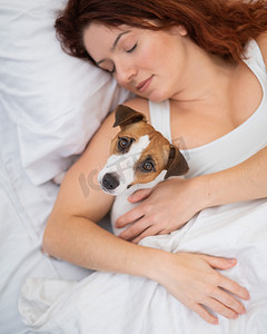 睡着的狗摄影照片_白人妇女抱着她心爱的狗在床上打盹。