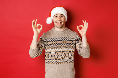 快乐而快乐的帅哥的画像，穿着冬衣和圣诞老人​​的帽子，表现出好的迹象，赞美圣诞晚会，站在红色背景上