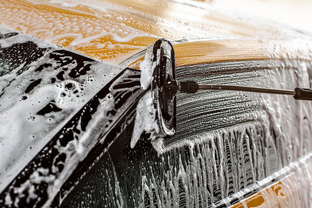 洗车请左转谢谢摄影照片_黄色汽车的侧面正在自助洗车中清洗，刷子在玻璃上留下了洗发水和泡沫的痕迹。
