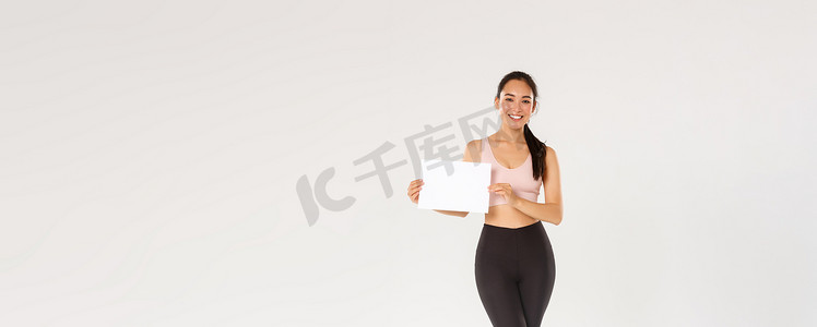 全长微笑的漂亮亚洲黑发女运动员、穿着运动服的女运动员在白纸上展示标志、广告健身房会员资格或健身器材特价