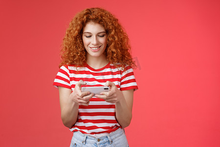 红头发的女孩在排队等候的牙医身边傻傻地玩着很棒的智能手机游戏拿着手机水平点击手机屏幕看电话显示微笑高兴娱乐站立红色背景