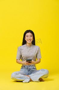 购物app摄影照片_温柔的亚洲年轻女孩坐在地板上，手里拿着手机发短信或网上购物，玩黄色背景的游戏。
