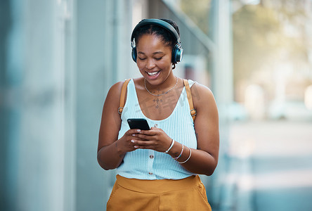 城市黑人女性戴着智能手机和耳机在城市或街道上行走、听音乐、联网并在聊天应用程序上打字。