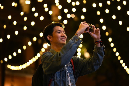 年轻时尚的男旅行者站在夜间街灯背景模糊的夜城里用相机拍照