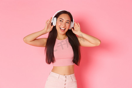时尚漂亮的亚洲女孩的肖像，戴着耳机享受听音乐，带着快乐的微笑，看起来梦幻而乐观，戴着无线耳机，站在粉红色的背景下